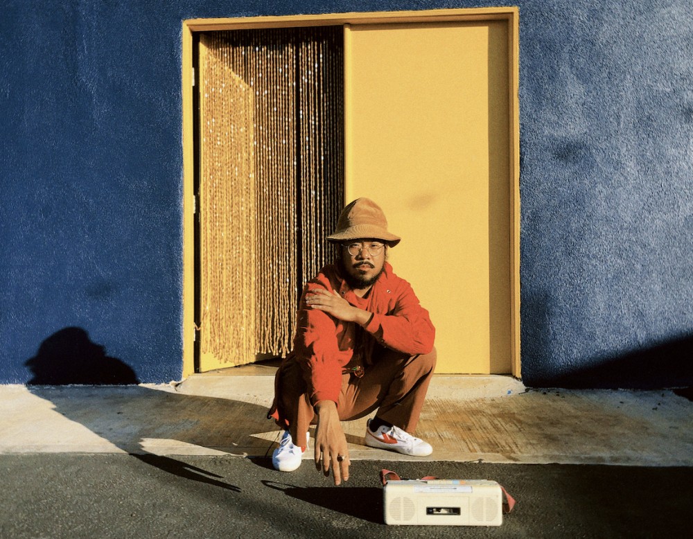 Musician and producer MNDSGN in burnt orange and khaki against a yellow door and cold blue wall.